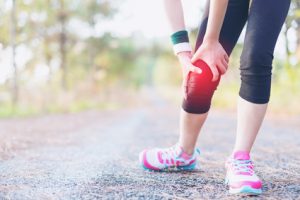 Eine Person in Sportkleidung steht auf einem Weg und hält sich mit beiden Händen das Knie, was auf Schmerzen oder Verletzungen hinweist.