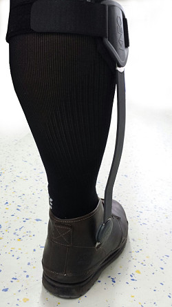 Person, die eine schwarze Orthese am Unterschenkel trägt, die über eine schwarze Socke und einen braunen Schuh gestülpt ist, steht auf einem gesprenkelten Boden.