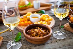 Zwei Gläser Weißwein auf einem Holztisch mit Schalen mit Käsewürfeln, Nüssen, Chutney und anderen Snacks.
