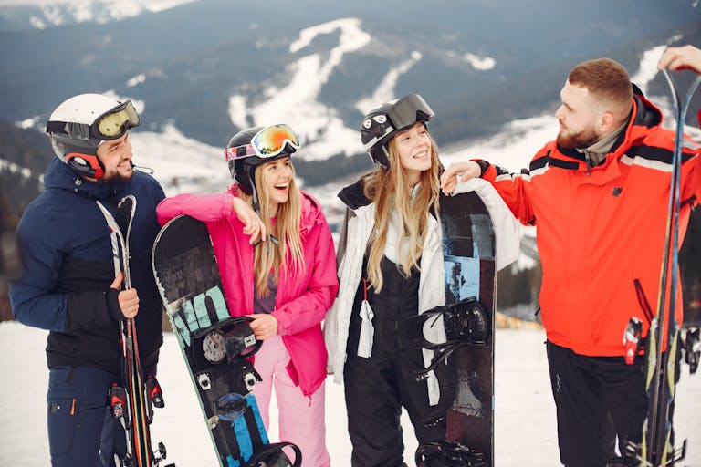 Four friends laughing and posing with snowboards and skis on a sunny winter day in snowy mountains.
