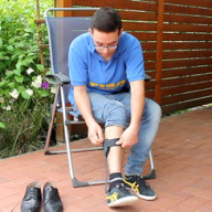 Ein Mann, der im Freien auf einem Stuhl sitzt, bindet sich ein schwarzes Band um den Knöchel, während seine Schuhe seitlich auf dem Boden liegen.