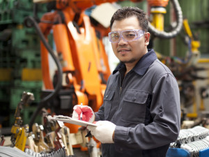 Ein Mann mit Schutzbrille und Handschuhen hält ein Klemmbrett in einer Fabrikumgebung mit Industrieanlagen im Hintergrund.
