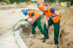 Zwei Bauarbeiter mit orangefarbenen Westen und Helmen heben einen Graben aus und verlegen Kabel neben einer im Bau befindlichen Straße.