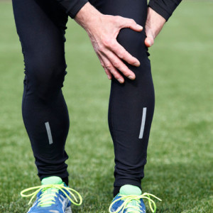 Eine Person, die schwarze Sportleggins und grüne Laufschuhe trägt, hält sich das Knie, was auf mögliche Knieschmerzen oder -verletzungen hindeutet, und steht auf einer Wiese.