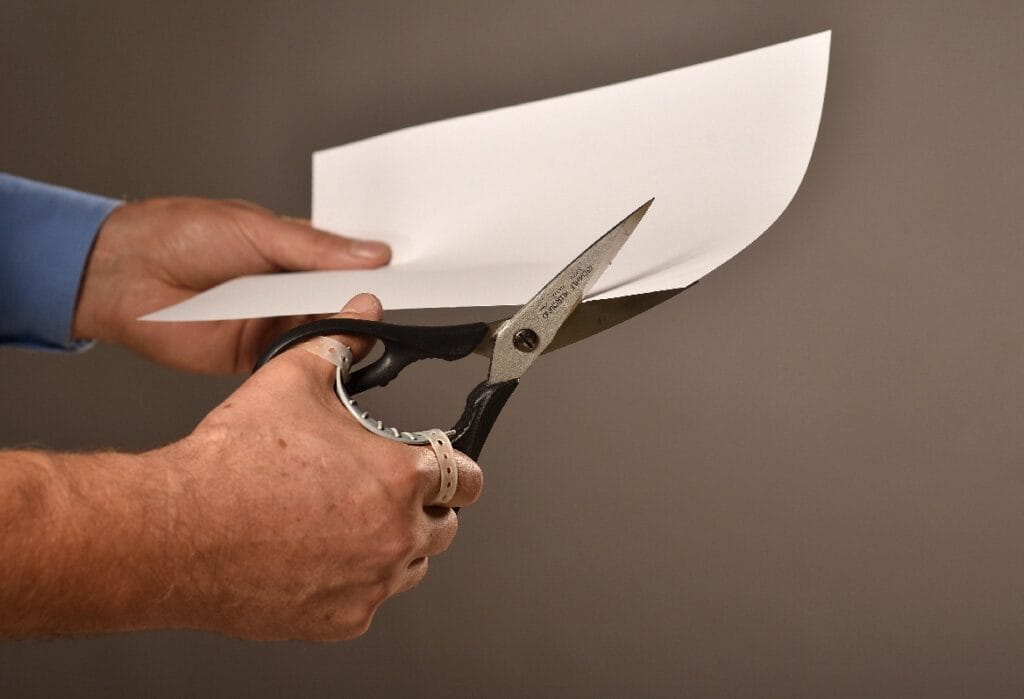 Eine Person benutzt eine Schere mit schwarzem Griff, um ein weißes Blatt Papier vor einem unifarbenen Hintergrund zu schneiden.
