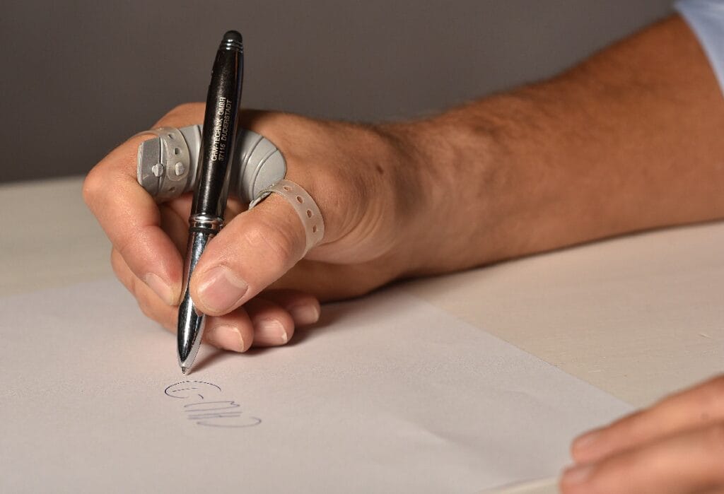 Eine Hand mit Verbänden und Schienen hält einen Stift und schreibt auf ein weißes Blatt Papier.