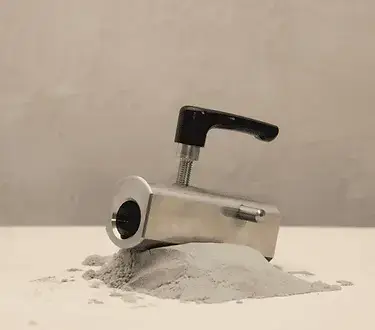 Ein Metallgerät mit schwarzem Griff ist teilweise in einen Haufen grauen Pulvers auf einer glatten Oberfläche mit neutralem Hintergrund eingebettet.