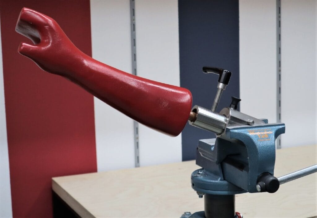 Eine rote Armprothese ist in einem blauen Metallschraubstock auf einer Werkbank befestigt, im Hintergrund ist eine gestreifte Wand zu sehen.