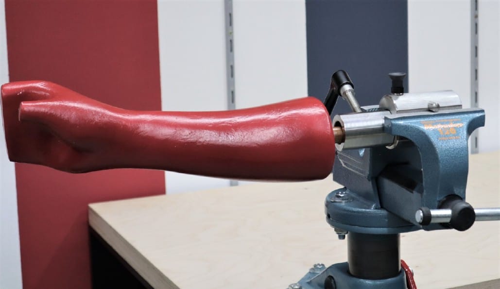 Ein rotes Armprothesenmodell ist in einem blauen Schraubstock auf einem Holztisch eingespannt, der mit einer gestreiften Wand hinterlegt ist.